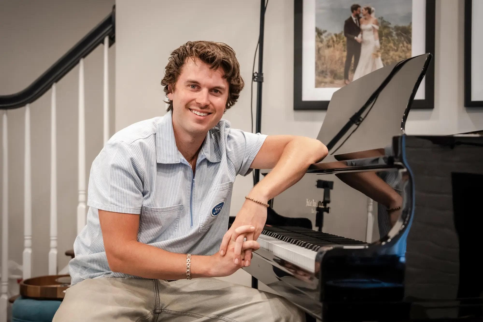 Photo of Cameron Francis smiling for the camera, while sitting on a piano bench and leaning against the baby grand piano in his parents' house